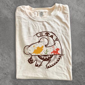 May include: A cream-colored short-sleeve t-shirt featuring a brown graphic print of three animal silhouettes, including a warthog, a meerkat, and a lion cub, framed by stylized line art of park landmarks and geometric patterns with small star accents.