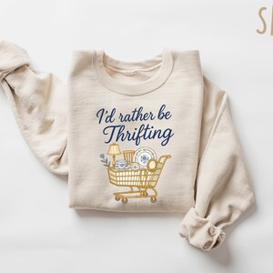 May include: Sand-colored sweatshirt with the text "I'd rather be thrifting" in navy blue script. A graphic of a shopping cart filled with vintage items is below the text. The sweatshirt is folded on a white surface.