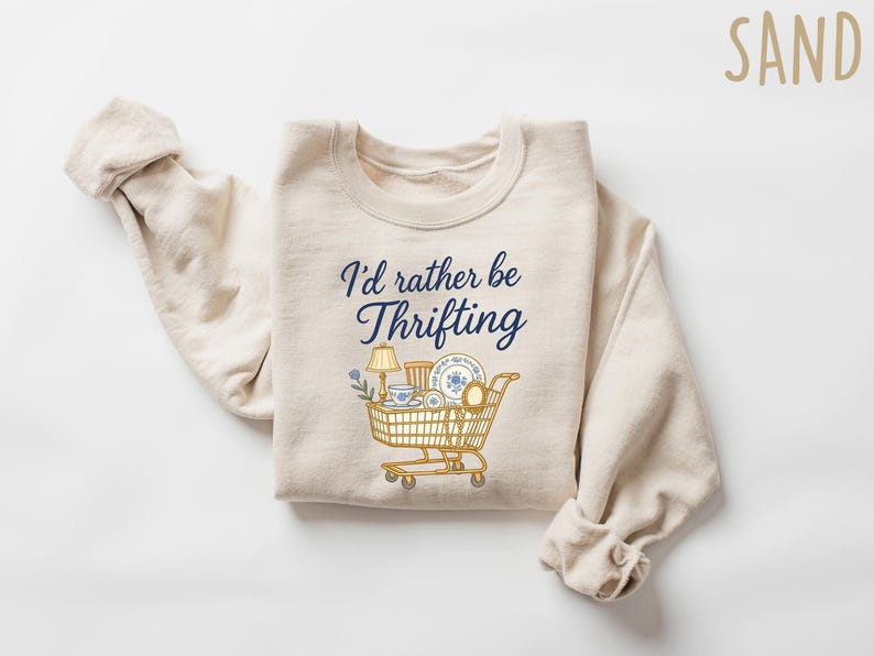 May include: Sand-colored sweatshirt with the text "I'd rather be thrifting" in navy blue script. A graphic of a shopping cart filled with vintage items is below the text. The sweatshirt is folded on a white surface.