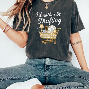May include: Dark gray t-shirt with the text "I'd rather be Thrifting" and a graphic of a shopping cart filled with thrifted items. The cart includes a lamp, dishes, and other vintage finds.