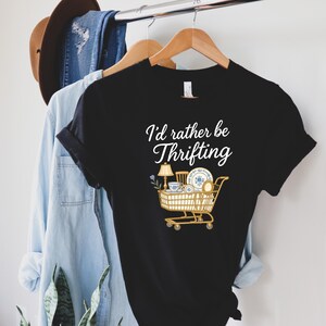 May include: Black t-shirt with the text "I'd rather be Thrifting" in white script. The shirt features a graphic of a shopping cart filled with vintage items, including a lamp, teacup, and plates.
