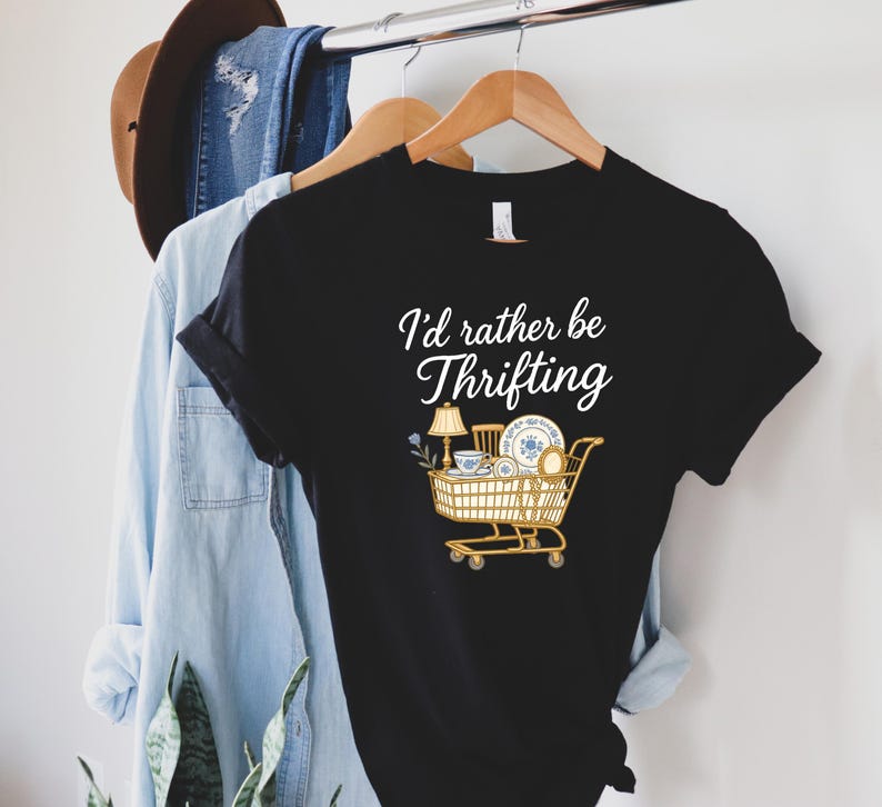 May include: Black t-shirt with the text "I'd rather be Thrifting" in white script. The shirt features a graphic of a shopping cart filled with vintage items, including a lamp, teacup, and plates.