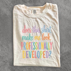 May include: A cream-colored short-sleeve t-shirt featuring the multicolored text does this shirt make me look PROFESSIONALLY DEVELOPED? The shirt has a crew neckline and a Comfort Colors brand label visible on the inside of the collar area.