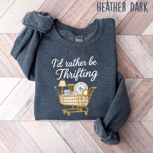 May include: Heather dark gray sweatshirt with the text "I'd rather be thrifting" in white script. A shopping cart graphic is filled with vintage items like a lamp, teacup, and plates. The sweatshirt is folded on a wooden surface.