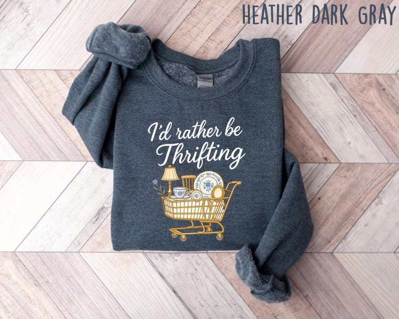 May include: Heather dark gray sweatshirt with the text "I'd rather be thrifting" in white script. A shopping cart graphic is filled with vintage items like a lamp, teacup, and plates. The sweatshirt is folded on a wooden surface.