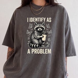 May include: A charcoal gray short-sleeve t-shirt featuring a graphic of a raccoon holding a coffee cup surrounded by butterflies. The text above the raccoon reads I IDENTIFY AS and the text below the raccoon reads A PROBLEM in a light cream color.