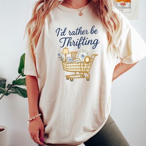 May include: Cream-colored t-shirt with the text "I'd rather be Thrifting" in navy blue script. A graphic of a shopping cart filled with vintage items like a lamp, dishes, and a mirror is below the text. The shirt is a casual, comfortable style.