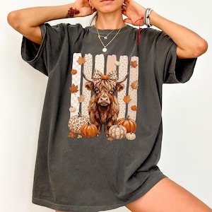 May include: A charcoal gray short-sleeve t-shirt featuring a graphic of a brown Highland cow wearing a leopard print bow. The cow is surrounded by orange and white pumpkins, scattered autumn leaves, and vertical striped panels in the background.
