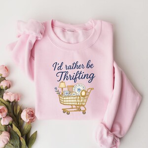 May include: A light pink sweatshirt with the phrase "I'd rather be Thrifting" in navy blue script. A shopping cart graphic is filled with vintage items. The sweatshirt is folded with a bouquet of pink flowers.