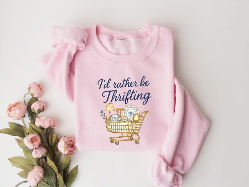 May include: A light pink sweatshirt with the phrase "I'd rather be Thrifting" in navy blue script. A shopping cart graphic is filled with vintage items. The sweatshirt is folded with a bouquet of pink flowers.