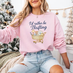 May include: A light pink sweatshirt with the text "I'd rather be thrifting" in navy blue script. A graphic of a shopping cart filled with thrifted items is below the text. The sweatshirt is a long-sleeved, crew neck style.