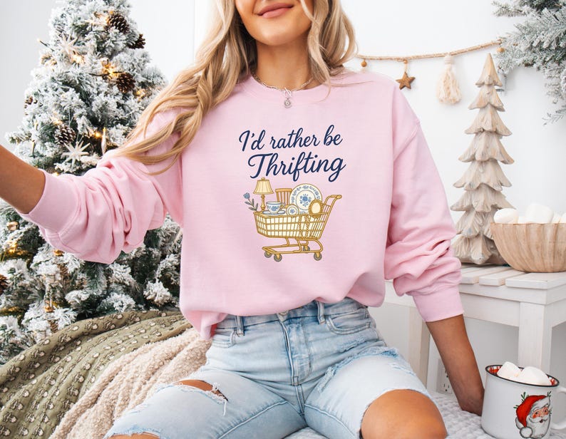 May include: A light pink sweatshirt with the text "I'd rather be thrifting" in navy blue script. A graphic of a shopping cart filled with thrifted items is below the text. The sweatshirt is a long-sleeved, crew neck style.