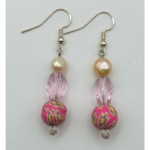 Könnte beinhalten: Ein Paar Ohrhänger mit silberfarbenen Haken, einer cremefarbenen Perle, einer facettierten, durchscheinenden rosa Perle und einer runden rosa Perle mit gelb-grünem Blumenmuster, abgeschlossen mit einem kleinen klaren Kristall am unteren Ende.