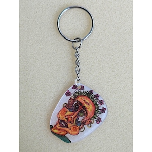 May include: A keychain with a silver ring and chain. The pendant is a clear, guitar pick-shaped design featuring a colorful, cartoonish face with flowers. The face is primarily orange, yellow, and green.