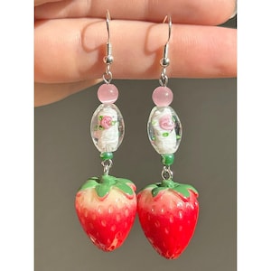 Könnte beinhalten: Ohrringe mit Erdbeermotiv. Jeder Ohrring hat einen rot-weißen Erdbeeranhänger, eine grüne Perle, eine ovale transparente Perle mit einer rosa Rose und eine rosa Perle. Die Ohrringe sind an silberfarbenen Haken befestigt.