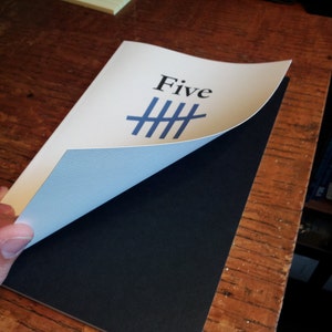 May include: A white card with the word "Five" printed in black and a blue tally mark symbol. The card is partially folded over a black surface.