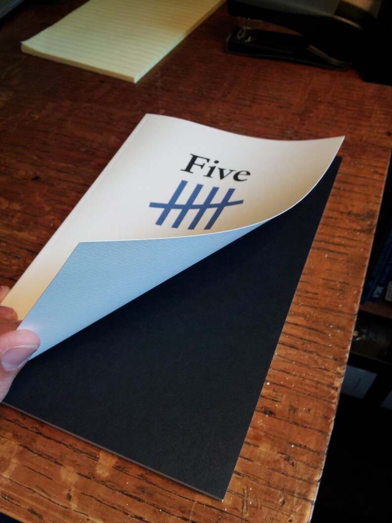 May include: A white card with the word "Five" printed in black and a blue tally mark symbol. The card is partially folded over a black surface.