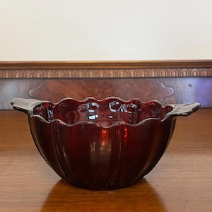 May include: A ruby red glass bowl with a scalloped rim and two small handles. The bowl has a ribbed design and sits on a wooden surface. The bowl is empty and reflects the light.