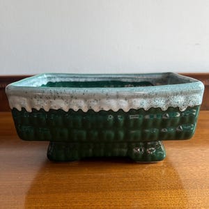 May include: A rectangular, ceramic planter with a dark green, textured exterior. The rim is a light gray with a white, speckled pattern. The planter sits on four small feet, and is suitable for small plants or succulents.