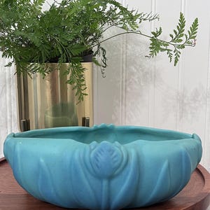 May include: A matte turquoise ceramic bowl featuring a molded floral motif around the exterior. The bowl has a scalloped rim and a rounded, low-profile shape. The surface has a smooth, non-reflective finish with subtle variations in color depth.