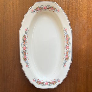 May include: An oval-shaped, off-white serving dish with a scalloped edge and a floral design. The dish has a silver trim and is decorated with pink roses, blue accents, and a brown ribbon pattern.