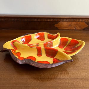 May include: A yellow ceramic serving dish with a leaf-like shape, featuring red and brown abstract designs. The dish has a dark brown rim and is placed on a wooden surface. This decorative piece is suitable for serving snacks or appetizers.