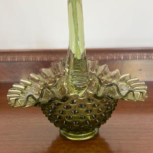 May include: A green glass basket with a handle. The basket has a ruffled rim and a textured, hobnail-like pattern on the body. The handle is arched and smooth, and the glass has a slight yellowish tint.