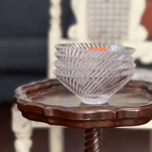 May include: A stack of clear glass bowls with a spiral design sits on a dark brown, ornate side table. The table has a carved, twisted leg and a scalloped edge. The bowls are stacked neatly, showcasing their textured pattern.