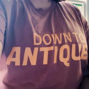 May include: A mauve t-shirt featuring the text "DOWN TO ANTIQUE" in orange. The crew neck shirt is made of a soft material. The image shows the top half of the shirt.