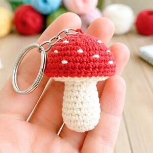 Pode incluir: Um chaveiro de cogumelo de crochê feito à mão. A tampa do cogumelo é vermelha com pontos brancos, e o caule é branco. O chaveiro tem um anel e uma corrente de metal prateado. O fundo inclui novelos de lã e um bloco de notas.