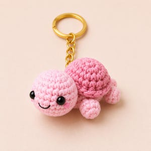 May include: A crochet turtle keychain featuring a light pink head with black plastic eyes and a stitched smile, attached to a gold-toned metal split ring. The turtle has a textured, darker pink shell and four small, rounded crochet legs.