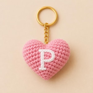 Puede incluir: Un llavero en forma de corazón hecho de hilo de ganchillo rosa con una letra P bordada en blanco en el centro. El corazón está unido a una anilla de metal dorado mediante un corto conector de cadena de metal dorado a juego.