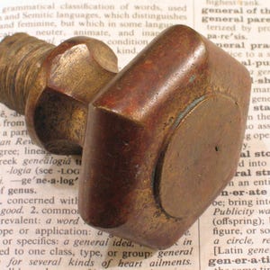 May include: A vintage, bronze-toned metal knob with a threaded base and a rectangular handle. The knob has a worn, aged appearance, suggesting it is an antique or a reclaimed piece.