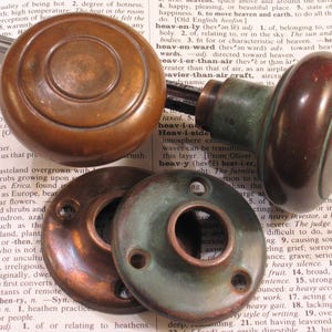 May include: Antique bronze door hardware set, including two doorknobs and two backplates. The doorknobs have a round shape with concentric circles, and the backplates are circular with three screw holes. The hardware has a vintage, aged appearance.