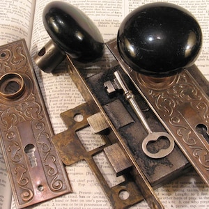 May include: Two black round door knobs with matching rectangular brass backplates featuring ornate embossed scrollwork patterns. A metal skeleton key and a brass mortise lock mechanism are positioned between the two door hardware sets.