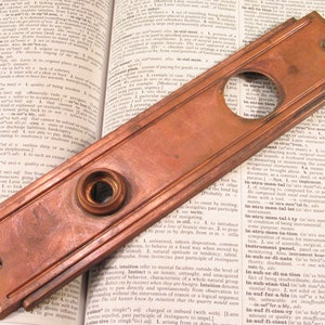 May include: A vintage copper door plate with a round hole and a smaller circular knob. The plate has a rectangular shape with a slightly raised edge and a smooth, polished surface.