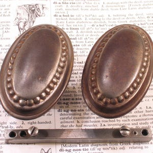 May include: Two antique bronze oval drawer pulls with a beaded edge and a matching metal bar with screw holes. These vintage hardware pieces are ideal for restoring furniture.