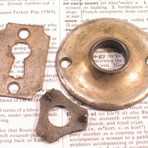 May include: Antique brass hardware components, including a keyhole plate, a circular door knob backplate, and a small, irregularly shaped piece. The pieces show signs of age and wear, with a vintage aesthetic.
