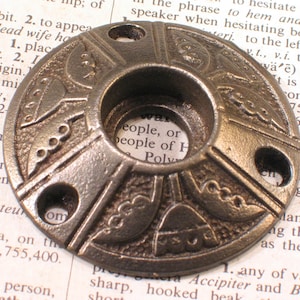 May include: A bronze-colored, circular metal object with a central hole and three smaller holes. The surface features an embossed, symmetrical design with stylized shapes and lines. The object appears to be a decorative fitting or hardware element.