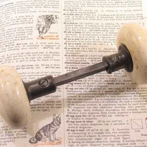 May include: An antique door handle with a cream-colored, oval-shaped grip and a dark metal stem. The handle is set against an open book with text and illustrations, suggesting a vintage or historical theme.