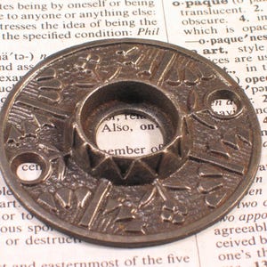 May include: A circular, dark bronze-toned metal escutcheon plate featuring a raised, textured floral and leaf relief pattern. The hardware component has a central circular opening surrounded by a serrated edge and two small screw holes.