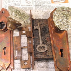 May include: Antique door hardware set featuring two ornate glass doorknobs, a key, and a lock mechanism. The metal components have a weathered, aged appearance with a copper-toned finish. The set is displayed on a vintage book page.