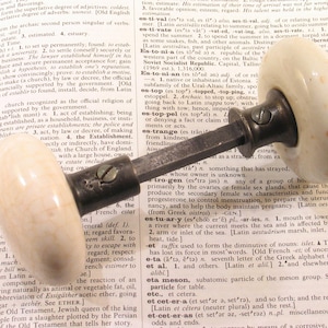 May include: An antique door knob with a cream-colored, speckled ceramic handle and a dark metal base. The knob is attached to a metal rod, resting on an open book with text.