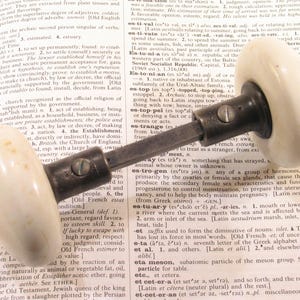 May include: An antique door knob with a cream-colored porcelain handle and a dark metal base. The knob is resting on an open book with text, suggesting a vintage or historical theme. The design is simple and classic.