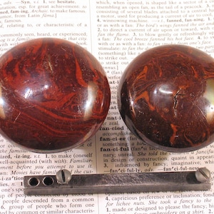 May include: Two antique drawer pulls or knobs with a swirled, reddish-brown pattern. The knobs are round and have a glossy finish. A metal bar with screws is also present, likely for installation. The items appear to be vintage.