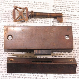 May include: Antique brass key and lock components. The key has an ornate handle. The lock parts include a rectangular plate with two holes and a smaller, rectangular piece. The items rest on an open book.