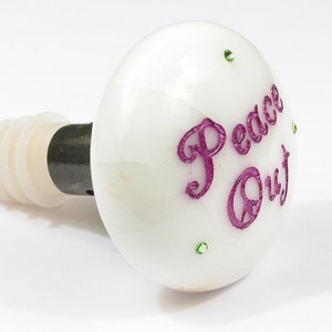 May include: White ceramic bottle stopper with a pink inscription that reads "Peace Out" and green glitter accents.