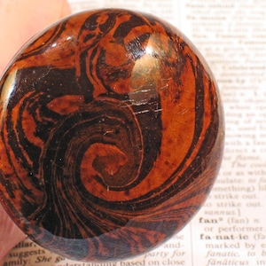 May include: A polished, round stone with a swirling pattern of dark brown and reddish-orange hues. The stone's surface is smooth and reflective, showcasing the natural veining and color variations.