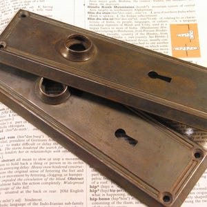 May include: Two rectangular door plates made of aged bronze metal. Each plate features a central circular knob collar, a keyhole opening, and four screw holes at the corners. The plates have a dark, weathered patina with visible metallic texture.