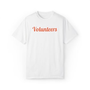 May include: A white short-sleeved t-shirt featuring the word Volunteers printed in a cursive, coral-colored font across the center of the chest. The garment has a standard crew neckline and a simple, clean design with short sleeves and a straight hem.